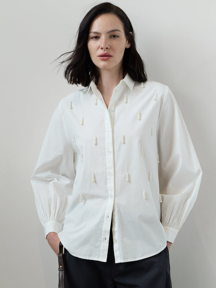 A woman wearing a white cotton Shirt with long balloon sleeves, a pointed collar, a button through fastening, faux pearl drop embellishments and a relaxed fit.