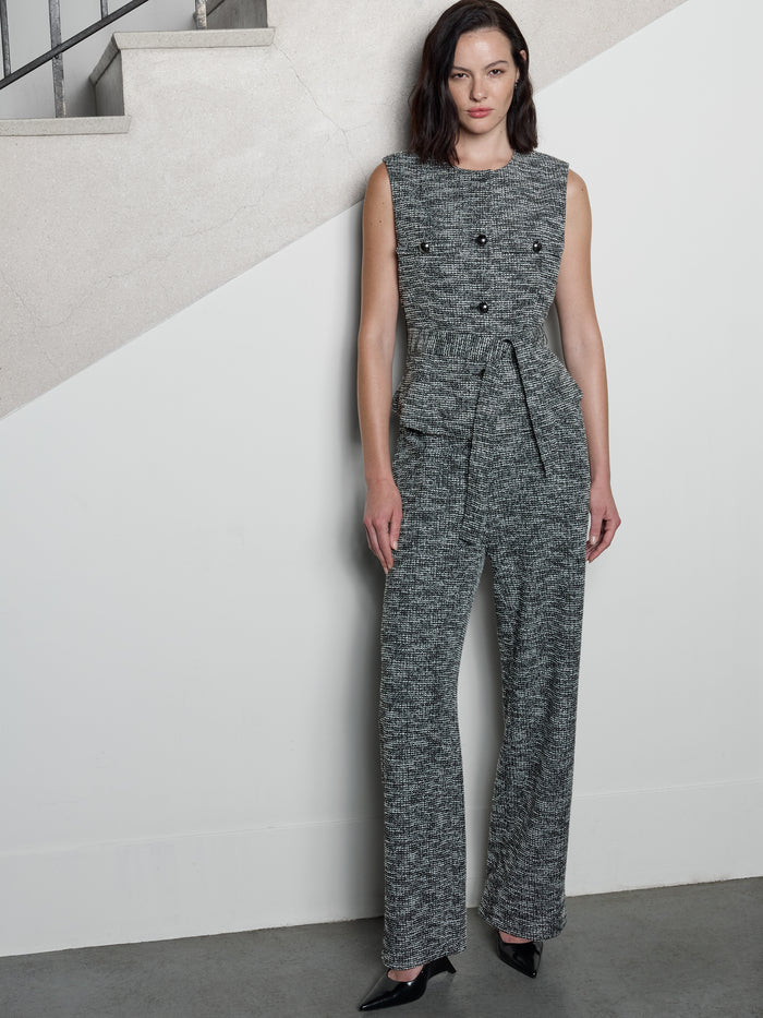 A woman wearing a tweed black and white sleeveless Waistcoat with a round neckline, metal button through fastening, 2 faux chest flap pockets and a self tie waist belt.